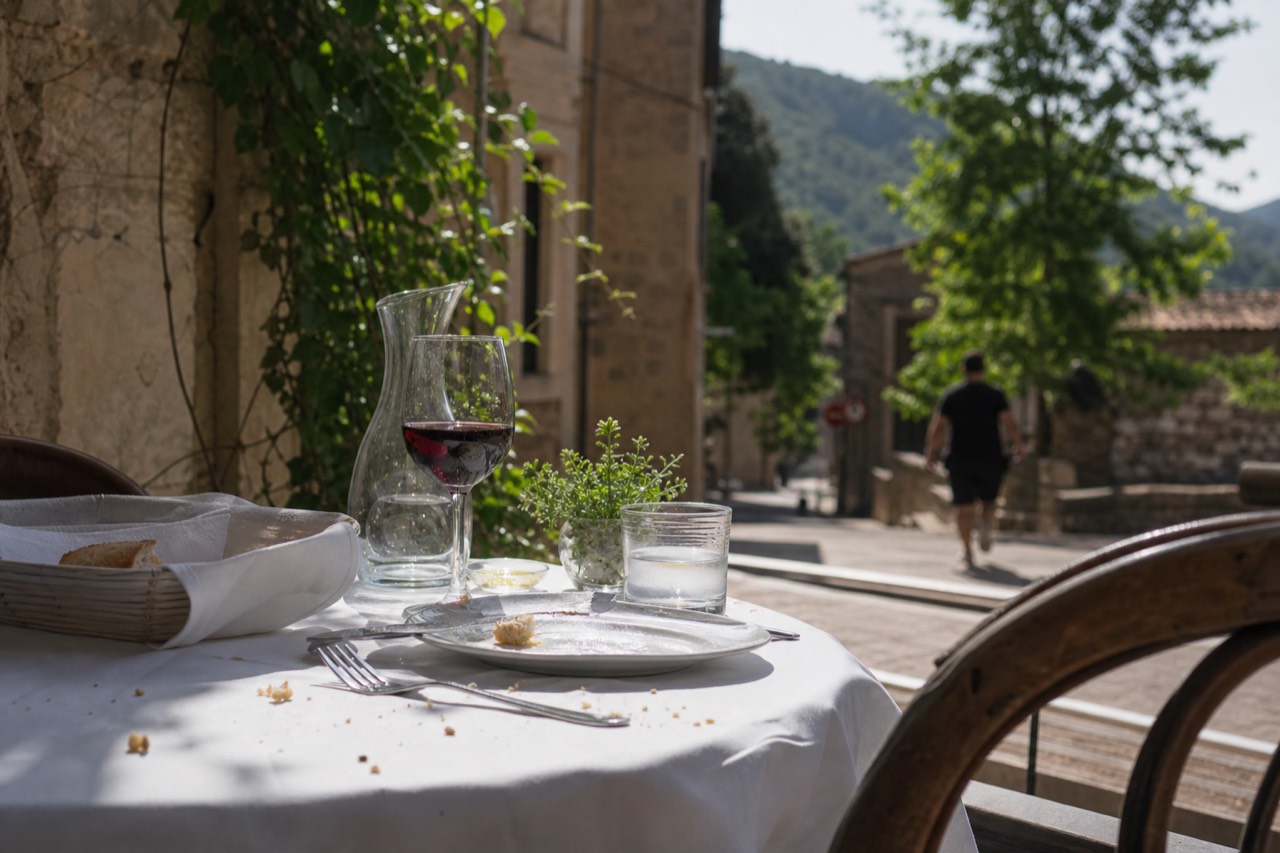 Sunlit terrace table set for lunch beside stone buildings in a medieval wine village, no text, no logos, no people.