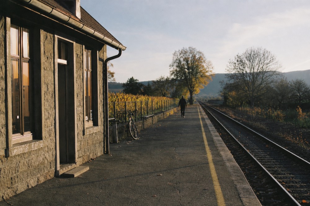 A small stone railway station beside vineyards at the edge of a village, no text, no logos, no people